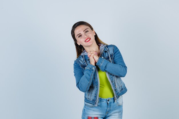 Young lady showing clasped hands in pleading gesture in denim outfit and looking jolly , front view.