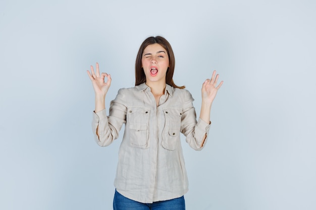 Young lady showing ok gesture while blinking in casual, jeans and looking glad. front view.