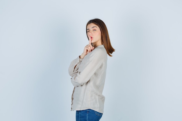 Young lady showing silence gesture while blinking in casual, jeans and looking cool. front view.