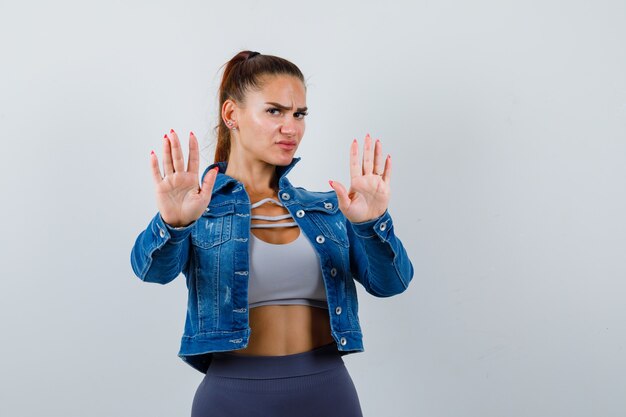 Young lady showing stop gesture in top, denim jacket and looking serious. front view.
