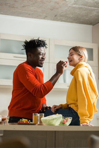 Giovane donna che sorride mentre il ragazzo premuroso positivo sta in piedi in cucina davanti a lei e la nutre con pomodorini