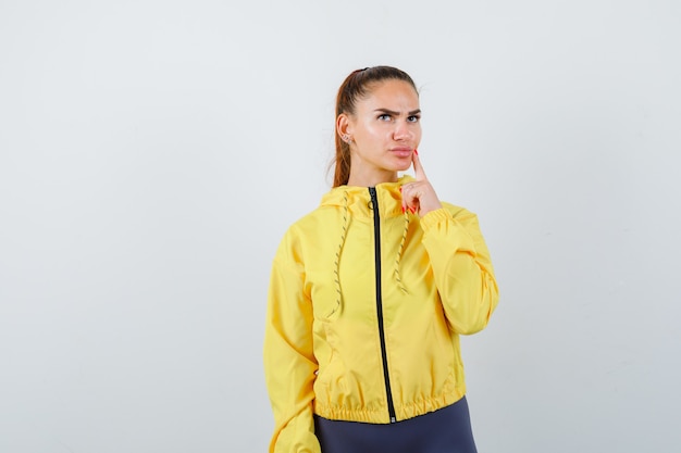 Young lady standing in thinking pose in yellow jacket and looking pensive , front view.