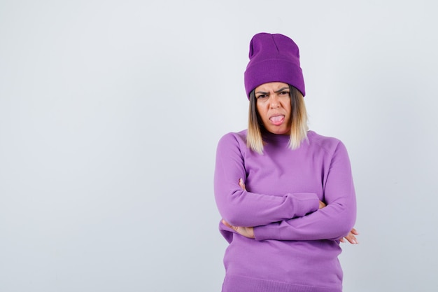 Young lady standing with crossed arms while sticking out tongue in purple sweater, beanie and looking resentful. front view.