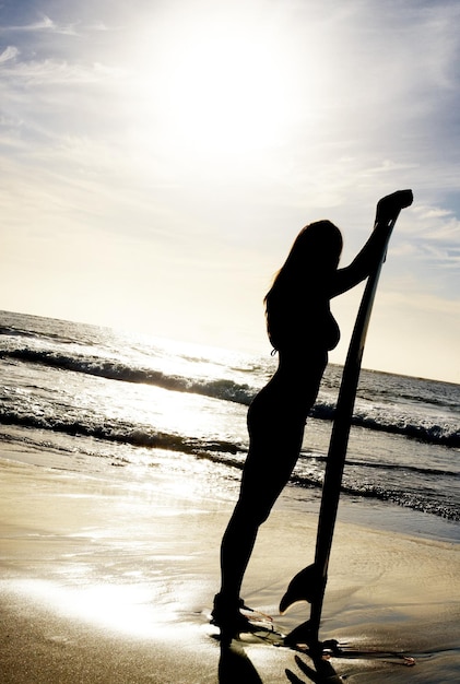 해질녘에 서핑보드와 함께 서 있는 젊은 여성 해변에서 서핑보드와 함께 서 있는 젊은 여성의 초상화