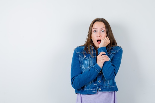 Young lady in t-shirt, jacket leaning cheek on hand and looking scared , front view.