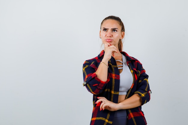 Giovane donna in alto, camicia a quadri con la mano sotto il mento e sguardo pensieroso, vista frontale.