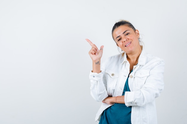 Young lady in white jacket pointing at upper left corner and looking cheery , front view.