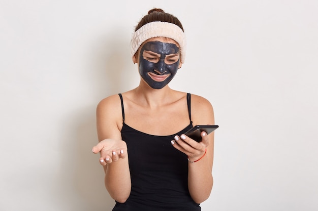 Young lady with black facial cosmetic mask using cellphone