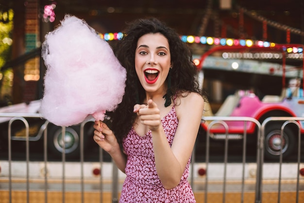 写真 ピンクの綿菓子を手に立って、遊園地のカメラで人差し指を見せながら幸せそうにカメラを見てドレスを着た暗い巻き毛の若い女性