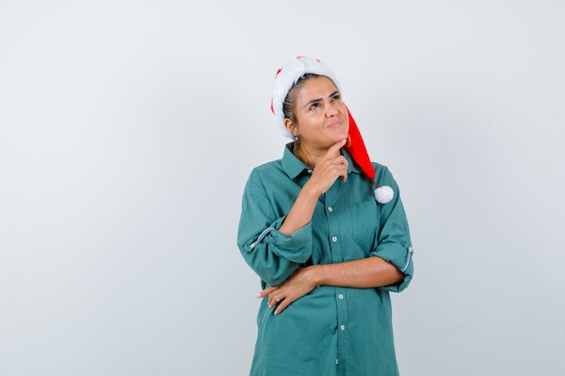 Giovane donna con il dito sotto il mento in cappello di natale, camicia e sguardo compiaciuto. vista frontale.