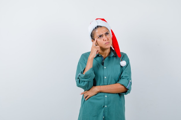 Young lady with finger on temples in christmas hat, shirt and looking pensive. front view.