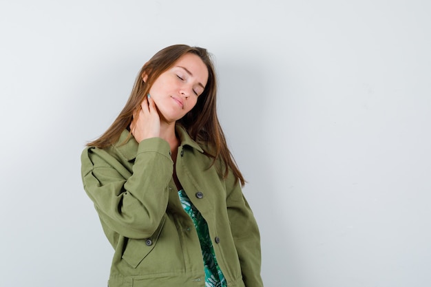 Young lady with hand on neck in green jacket and looking fatigued , front view.