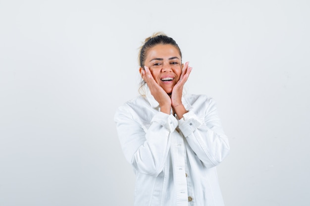 Young lady with hands on cheeks in shirt, white jacket and looking joyful. front view.