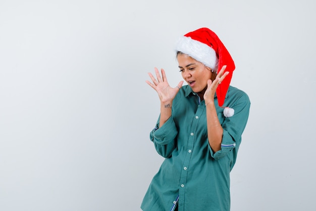 Giovane donna con le mani vicino al viso con cappello di natale, camicia e sguardo sorpreso. vista frontale.