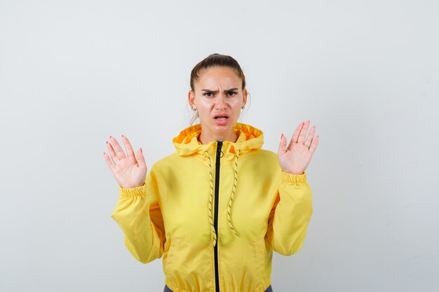 Young lady in yellow jacket keeping hands in surrender gesture and looking anxious , front view.