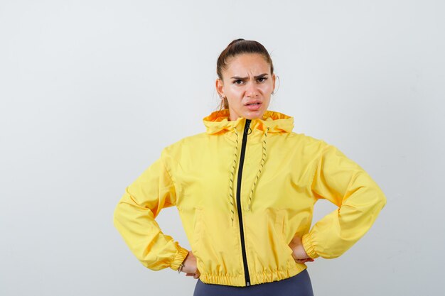 Young lady in yellow jacket with hands on waist and looking angry , front view.