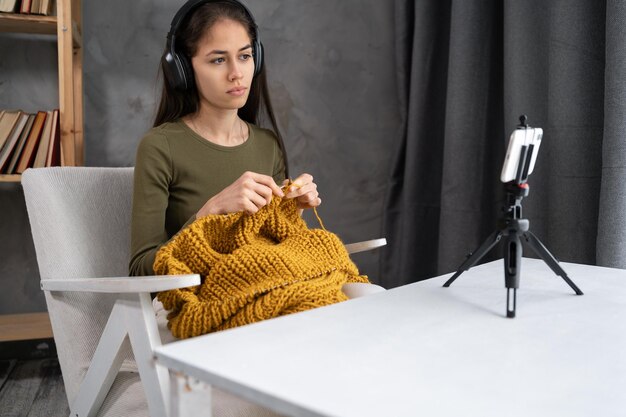 young latin blogger or designer shooting or videographing own needle knitting by using a smartphone camera on a tripod and a mobile phone for remote control technology and hobby
