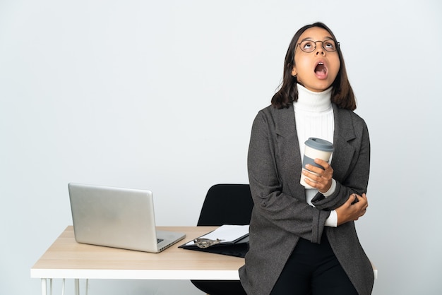 Молодая латинская деловая женщина, работающая в офисе, изолированном на белом фоне, смотрит вверх и с удивленным выражением лица
