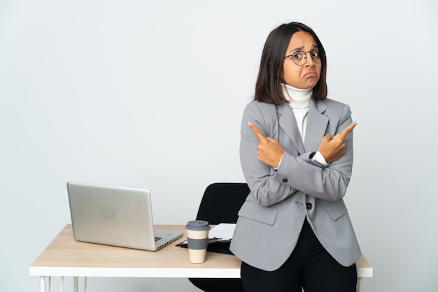 흰색 배경에 격리된 사무실에서 일하는 젊은 라틴 비즈니스 여성이 의심스러운 측면을 가리키는