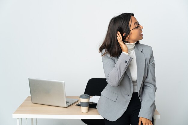 Giovane donna d'affari latino che lavora in un ufficio isolato su bianco ascoltando qualcosa mettendo la mano sull'orecchio