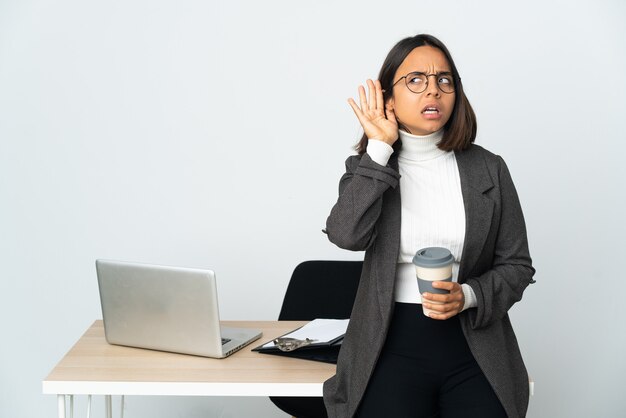 耳に手を置いて何かを聞いて白い壁に隔離されたオフィスで働く若いラテンビジネス女性