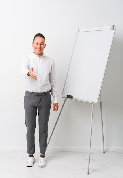 Young latin coaching man with a whiteboard isolated stretching hand in greeting gesture.