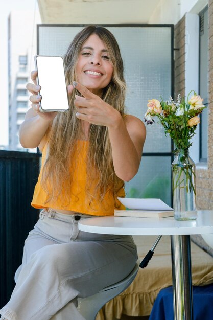 Young latin colombian woman with outstretched hand indoors showing her phone with blank screen