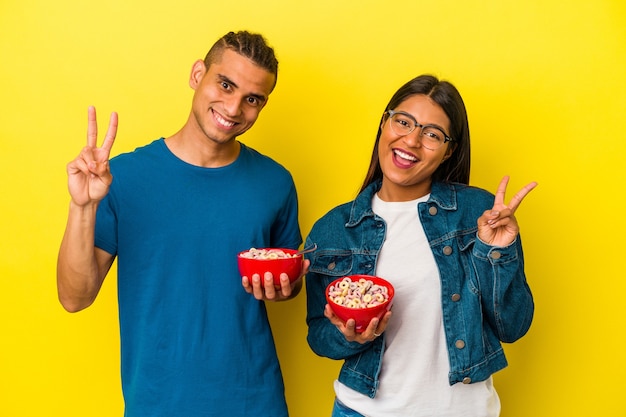 Young latin couple holding a cereals bowl isolated on yellow background joyful and carefree showing a peace symbol with fingers.