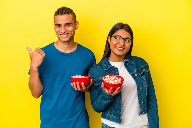 Young latin couple holding a cereals bowl isolated on yellow background points with thumb finger away, laughing and carefree.