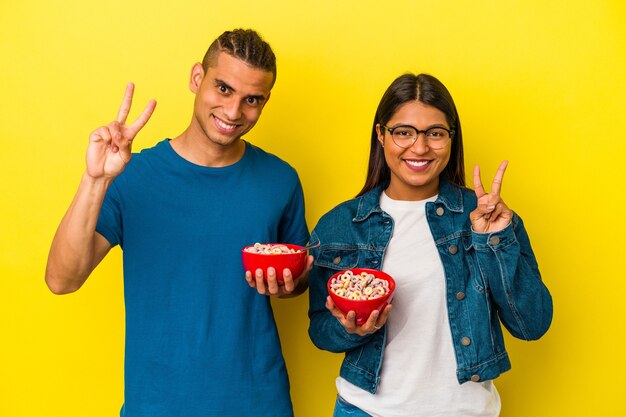 Young latin couple holding a cereals bowl isolated on yellow background showing number two with fingers.