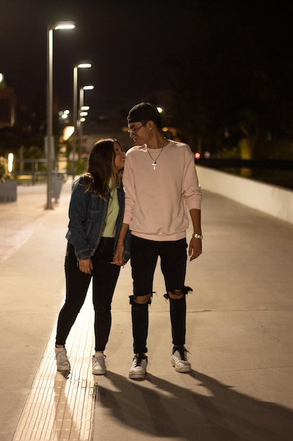 Young latin couple walking in the street at night holding\
hands, looking at each other with love, illuminated by the city\
lights and wearing urban style clothes.