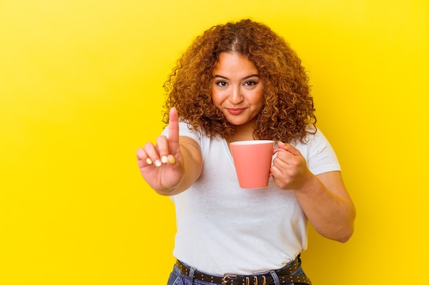 Young latin curvy woman holding a cup isolated on yellow background showing number one with finger.