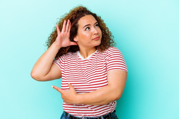 Foto giovane donna curvy latina isolata su priorità bassa blu che prova ad ascoltare un pettegolezzo.