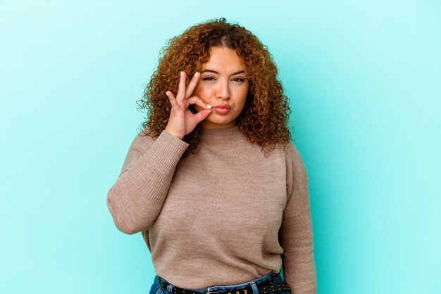 Young latin curvy woman isolated on blue background with fingers on lips keeping a secret.