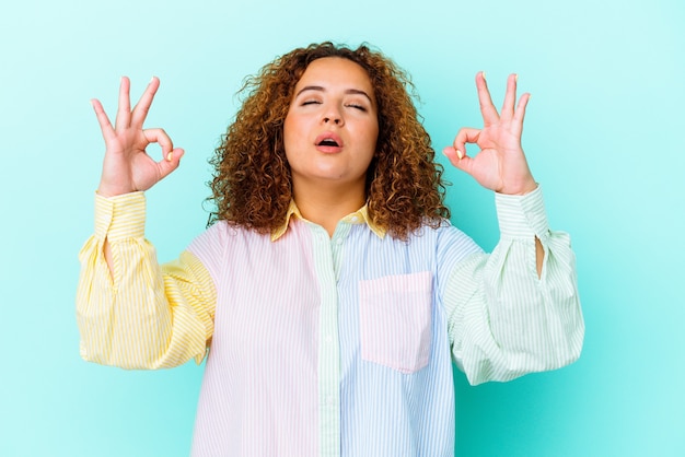 Young latin curvy woman isolated on blue wall relaxes after hard working day, she is performing yoga.