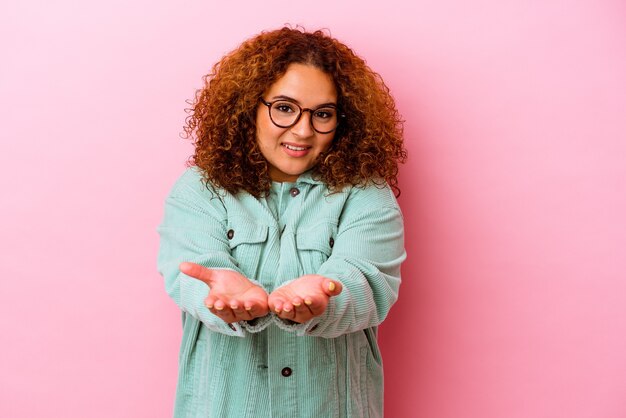 Young latin curvy woman isolated on pink background holding something with palms, offering to camera.