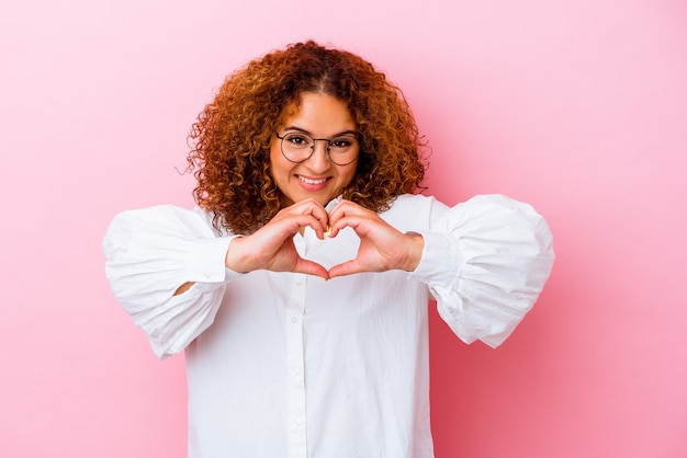 Young latin curvy woman isolated on pink background smiling and showing a heart shape with hands.