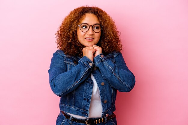 Young latin curvy woman isolated on pink wall keeps hands under chin, is looking happily aside.