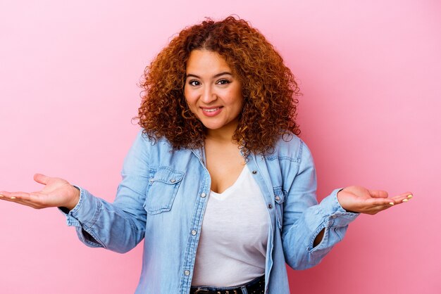 young latin curvy woman isolated on pink wall makes scale with arms, feels happy and confident.