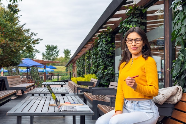 Young Latin female student wearing a yellow sweater holding a pen and looking straight at the camera in front of her laptop at a table outside