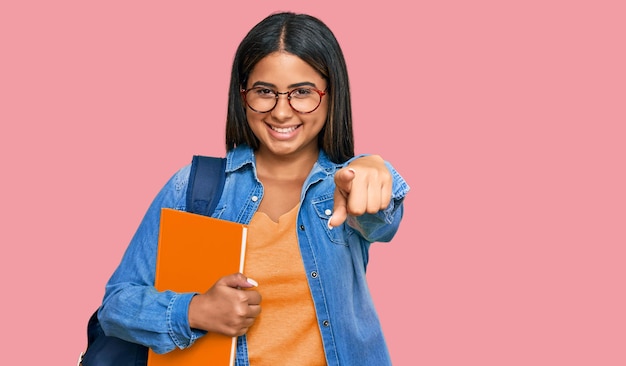 Young latin girl wearing student backpack and holding books pointing to you and the camera with fingers smiling positive and cheerful