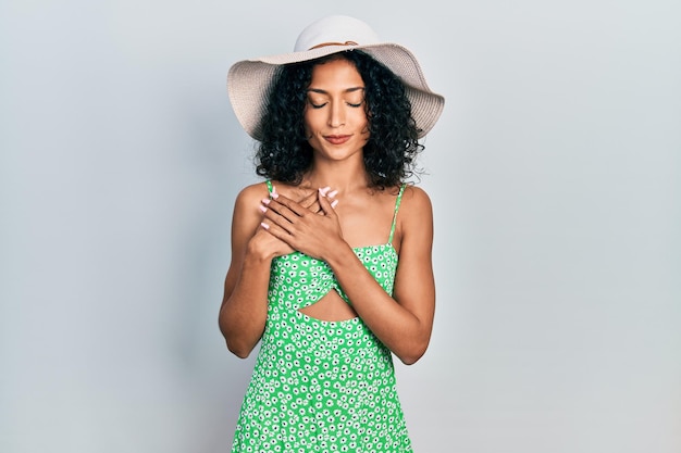 Young latin girl wearing summer hat smiling with hands on chest with closed eyes and grateful gesture on face. health concept.