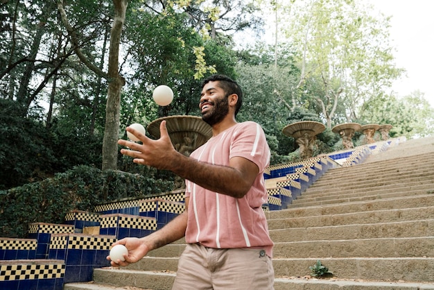 Young latin guy juggling in the sunny streets of Barcelona Doing a show with some white balls while smiling Dressed in brown pants and a pastel striped tshirt