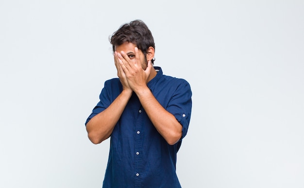 Young latin handsome man feeling scared or embarrassed, peeking or spying with eyes half-covered with hands