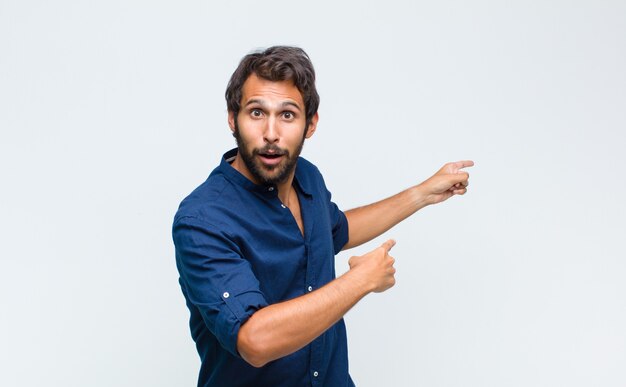 Young latin handsome man feeling shocked and surprised, pointing to copy space on the side with amazed, open-mouthed look