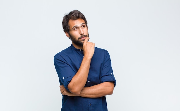 Young latin handsome man feeling thoughtful, wondering or imagining ideas, daydreaming and looking up to copy space