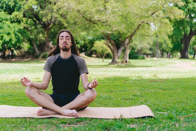 Young latin and hispanic hippie man with beard and long hair\
exhaling with eyes closed in a green field doing lotus pose on a\
yoga mat