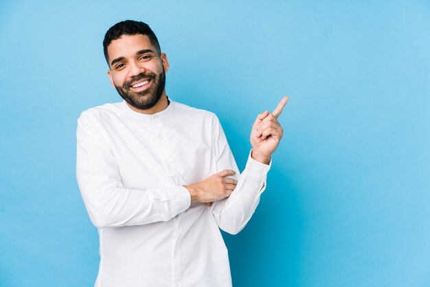 Il giovane uomo latino contro una parete blu ha isolato sorridere allegramente indicando con l'indice via.