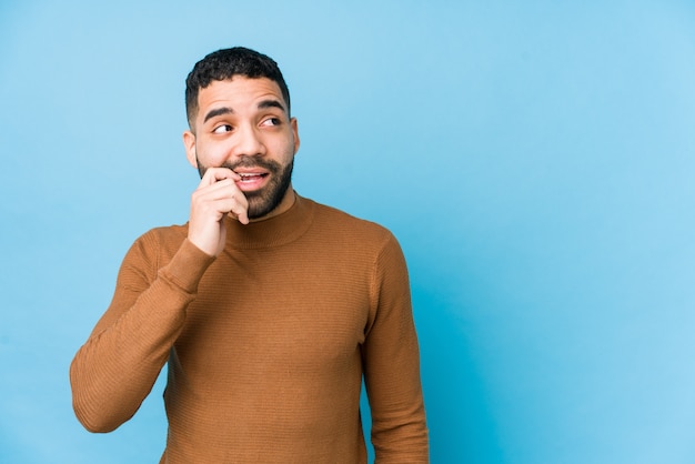 Young latin man against a blue wall relaxed thinking about something looking at a blank space.