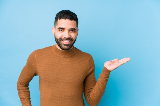 Young latin man against a blue wall showing a blank space on a palm and holding another hand on waist.